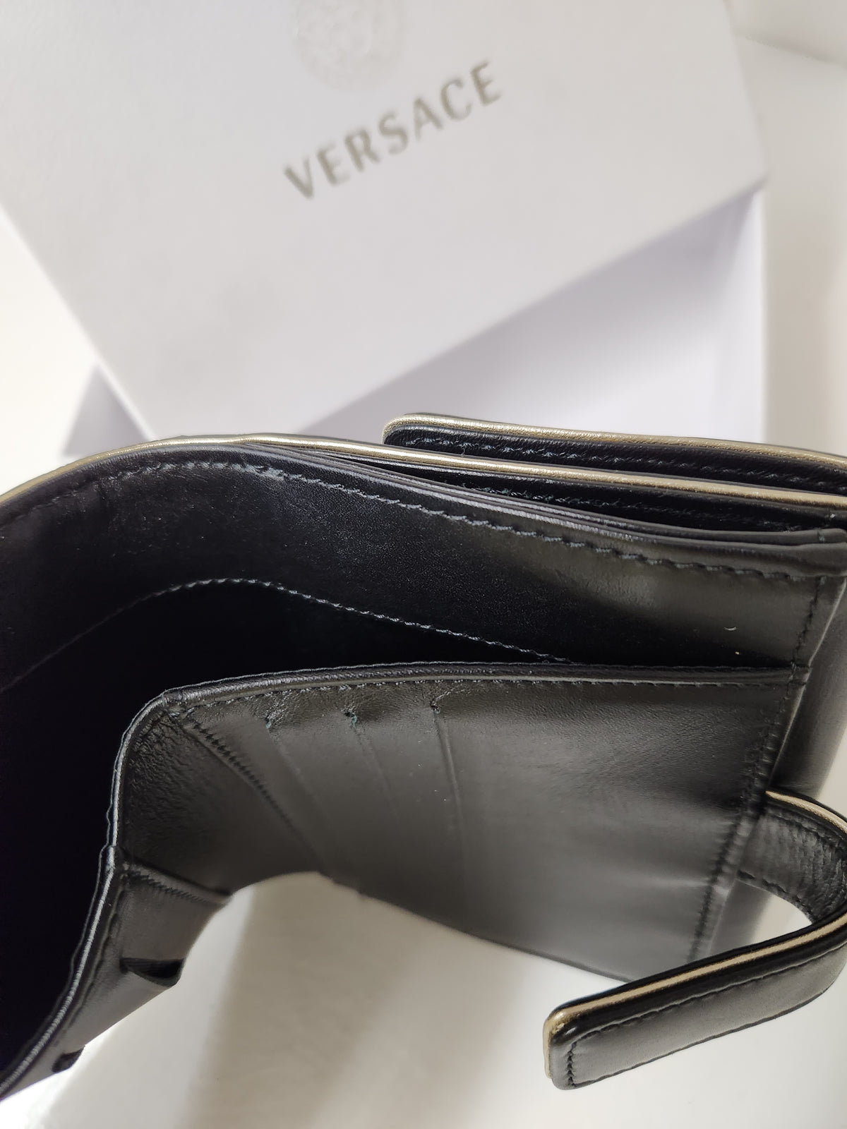 A close-up of a black leather Versace wallet or card holder, shown open with the interior and stitching visible, with the white Versace box in the background.