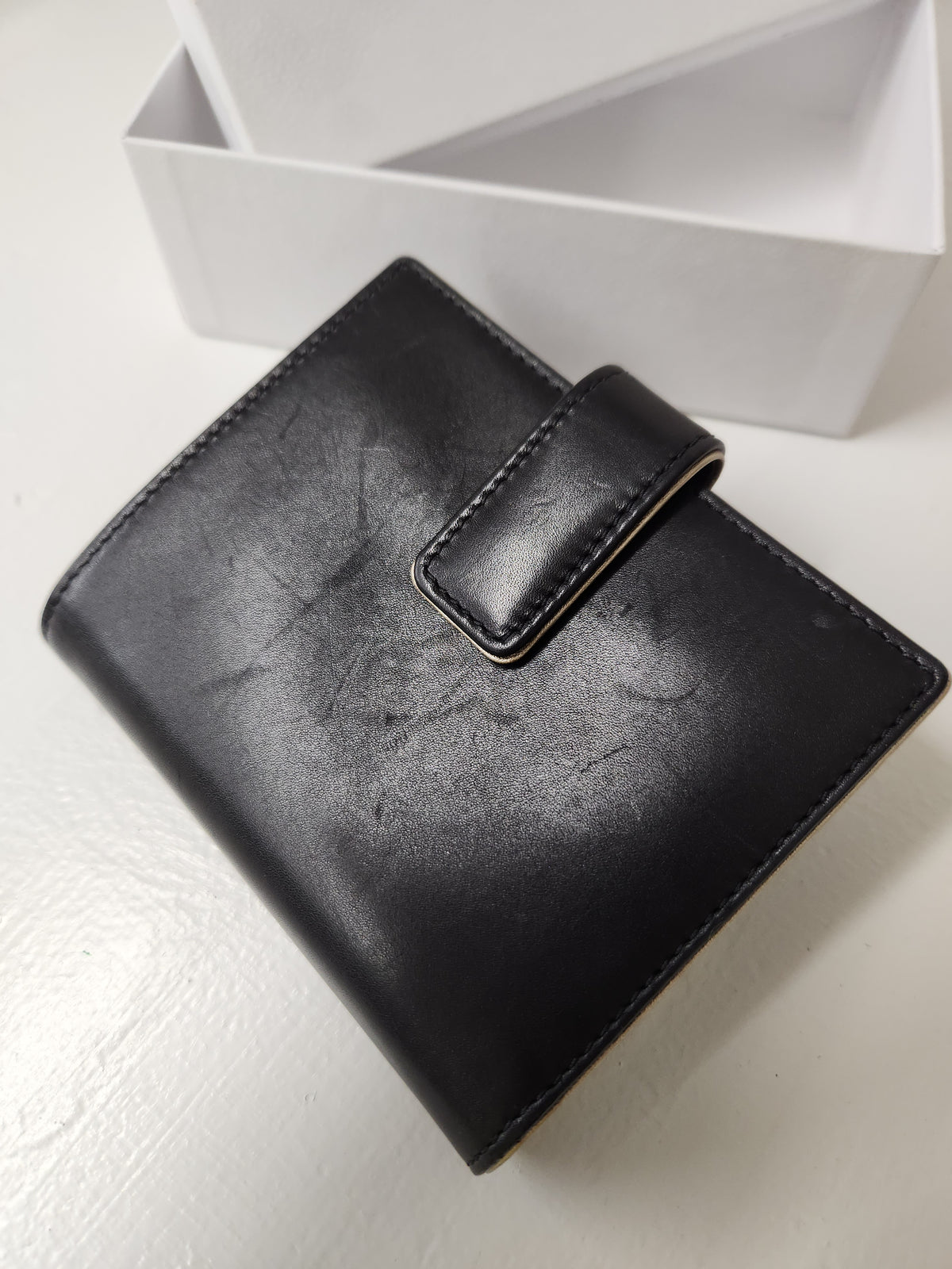 A black leather wallet with a strap closure, shown on a white surface with a white box in the background.