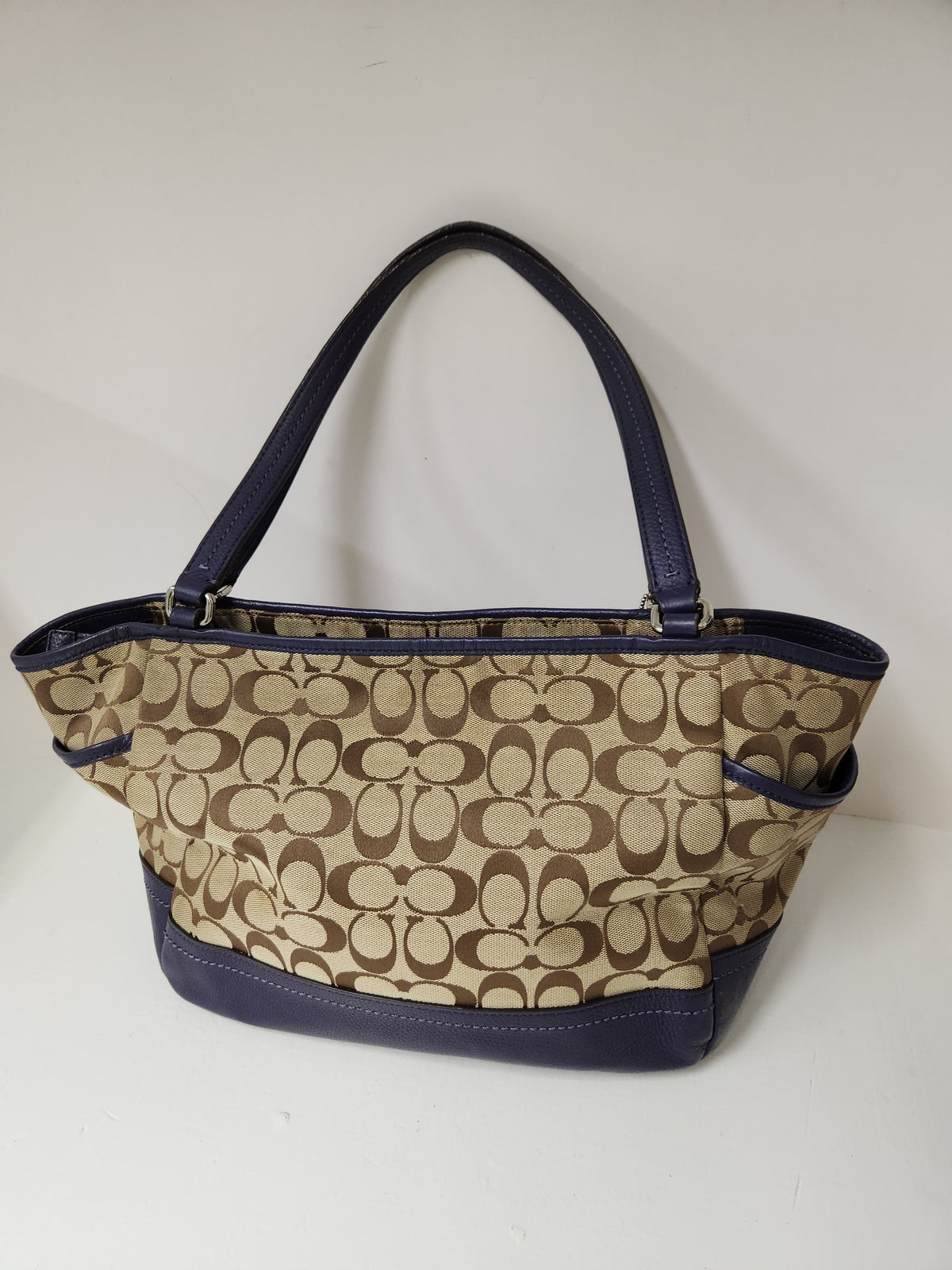 A Coach handbag in the classic signature monogram canvas, featuring a beige and brown pattern with navy blue leather trim and handles.
