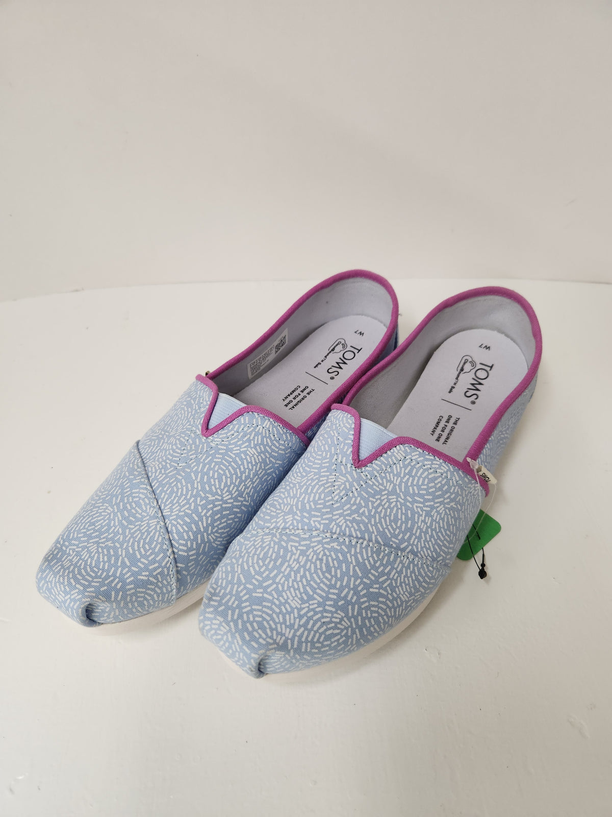 A pair of light blue canvas slip-on shoes with a white wave pattern, trimmed with pink piping and featuring a white insole with black branding.