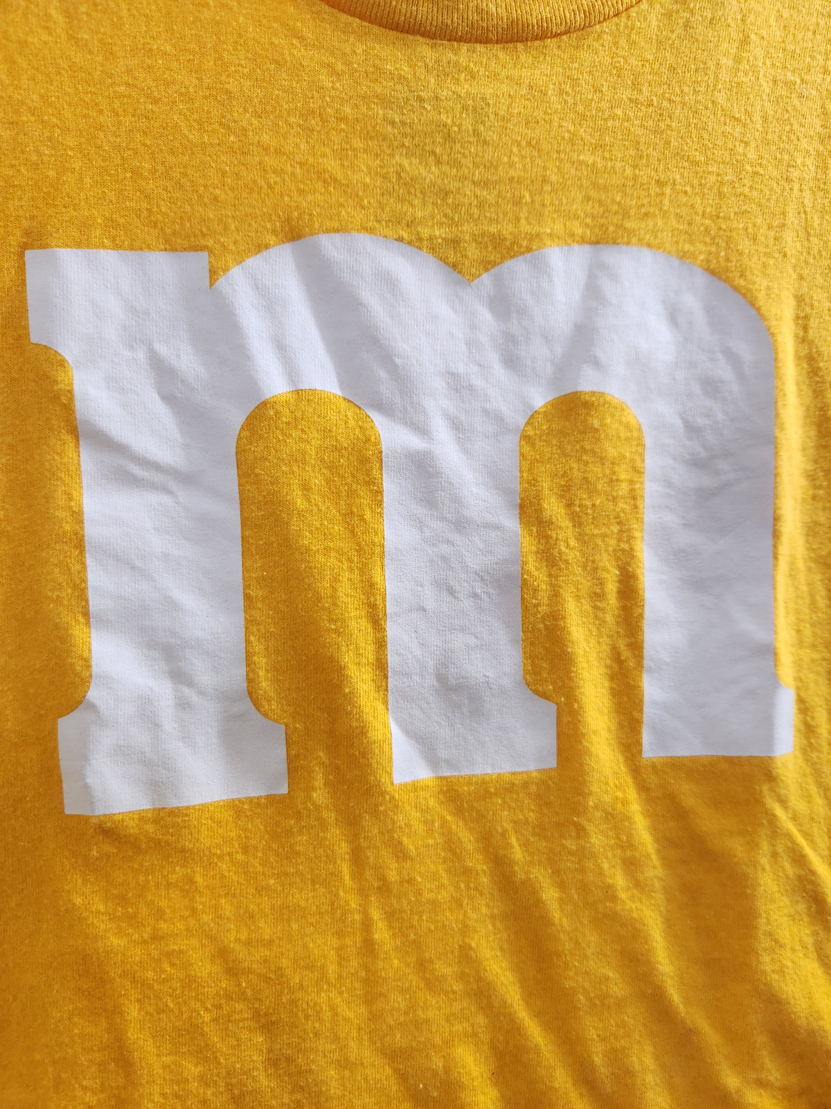 Close-up of a yellow t-shirt featuring a large white graphic letter 'm' resembling the M&M's logo.