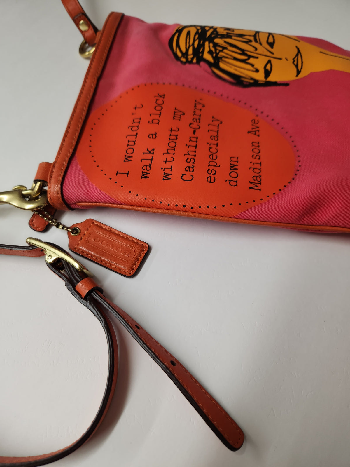 A pink and orange Coors-branded wristlet clutch with a brown leather strap and gold-tone hardware, featuring a stylized face illustration and a quote on a white background.