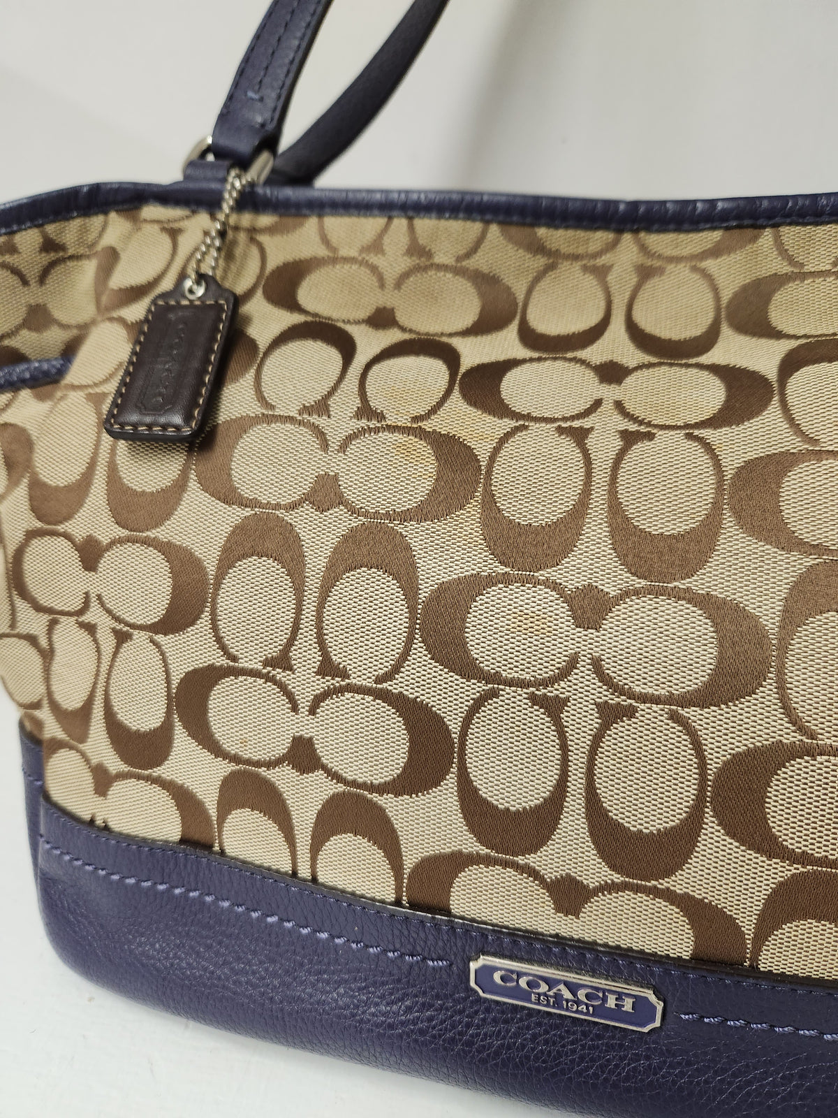 A close-up of a Coach handbag featuring the signature brown and beige monogram canvas body with navy blue leather trim and handles. A small metal logo plate is visible on the bottom right, and a leather hangtag is attached on the left.