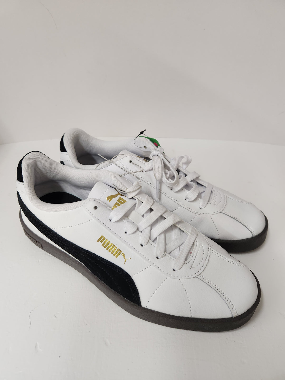 A pair of white Puma sneakers with black side stripes and gold branding, photographed on a light gray surface.