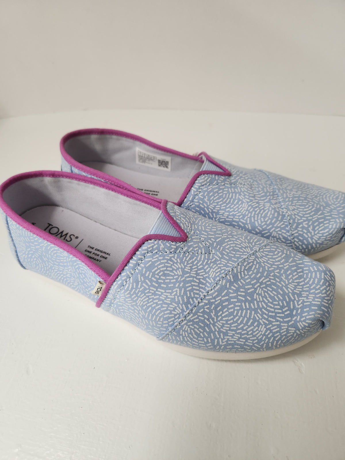 A pair of TOMS slip-on canvas shoes in light blue with a white textured pattern, featuring magenta-pink piping and white soles.