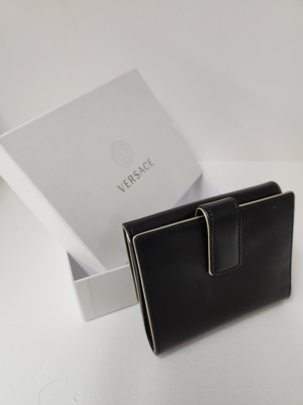 A black leather Versace wallet with gold trim, shown next to its white branded presentation box on a light gray background.