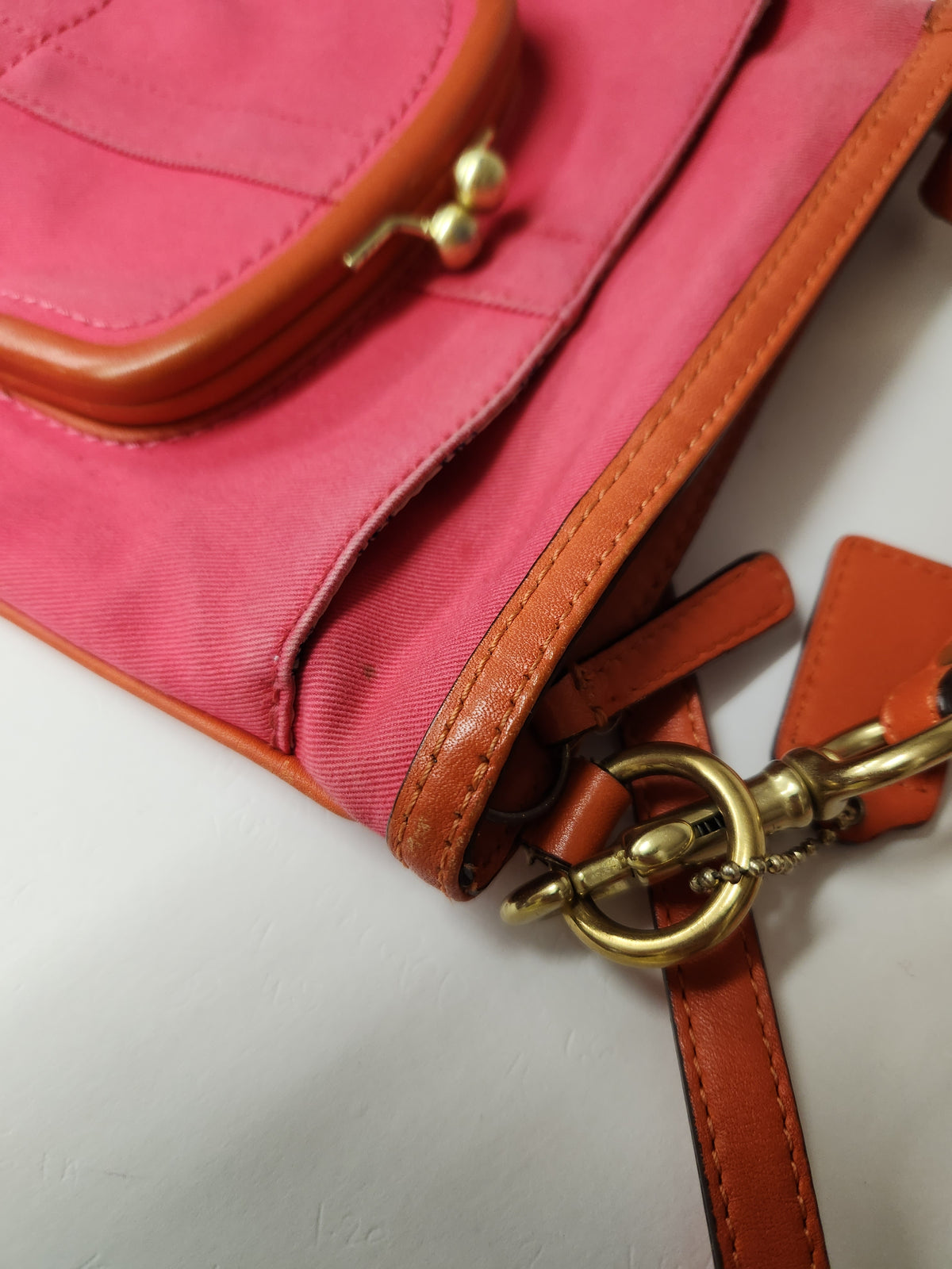 A close-up of a pink fabric handbag with tan leather trim and gold-tone hardware, including a clasp and ring detail.