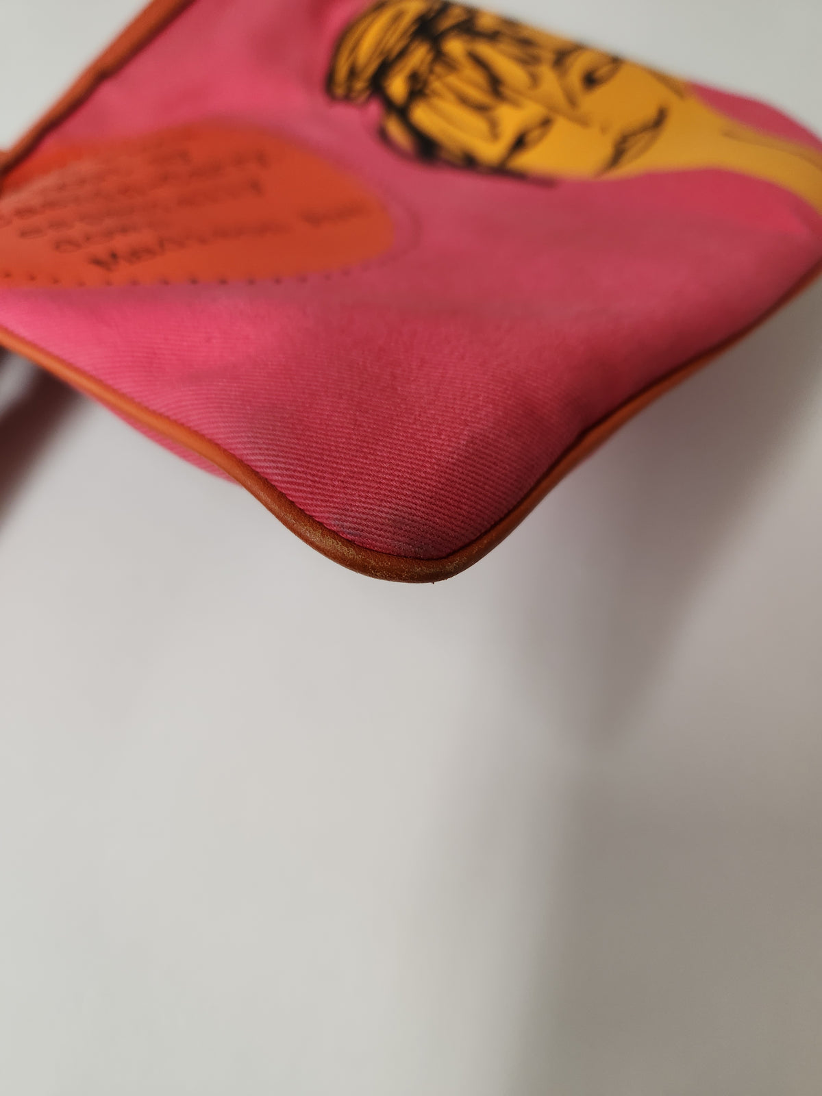 A close-up of a pink fabric pouch or small bag with a yellow illustrated face and an orange heart-shaped patch, photographed on a plain light background.