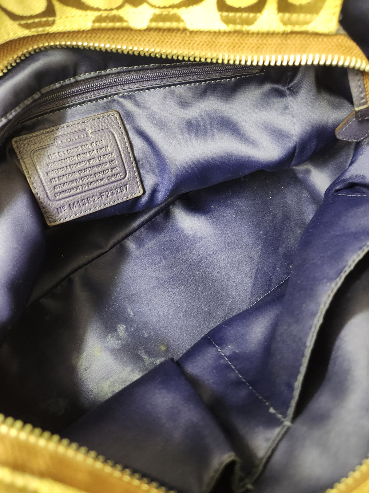 Close-up of the interior of a gold and navy blue bag, showing a dark blue satin lining, a zippered pocket, and a leather brand patch that reads Coach