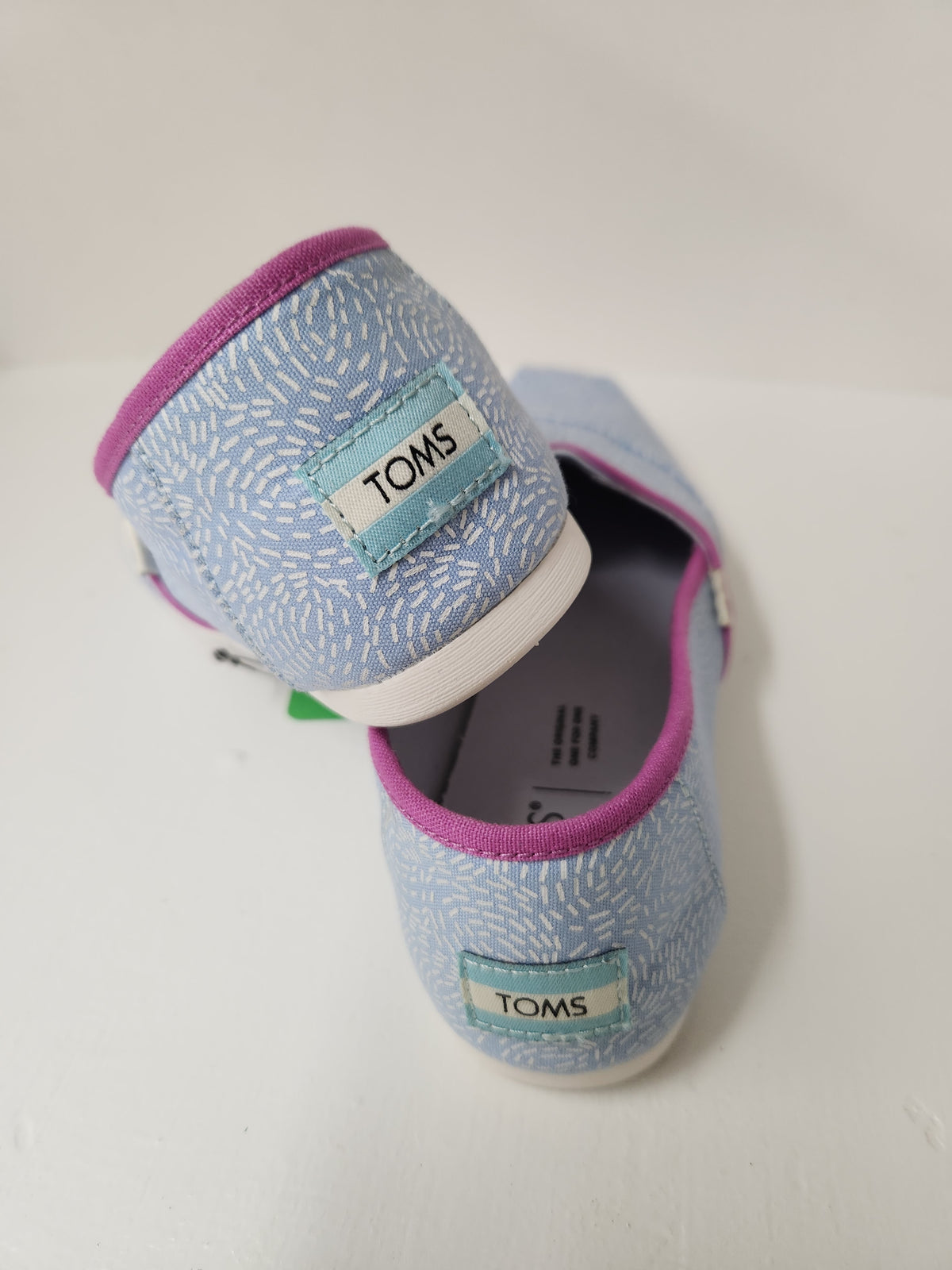 A pair of TOMS slip-on canvas shoes in light blue with a white brushstroke pattern, featuring magenta trim and white soles.
