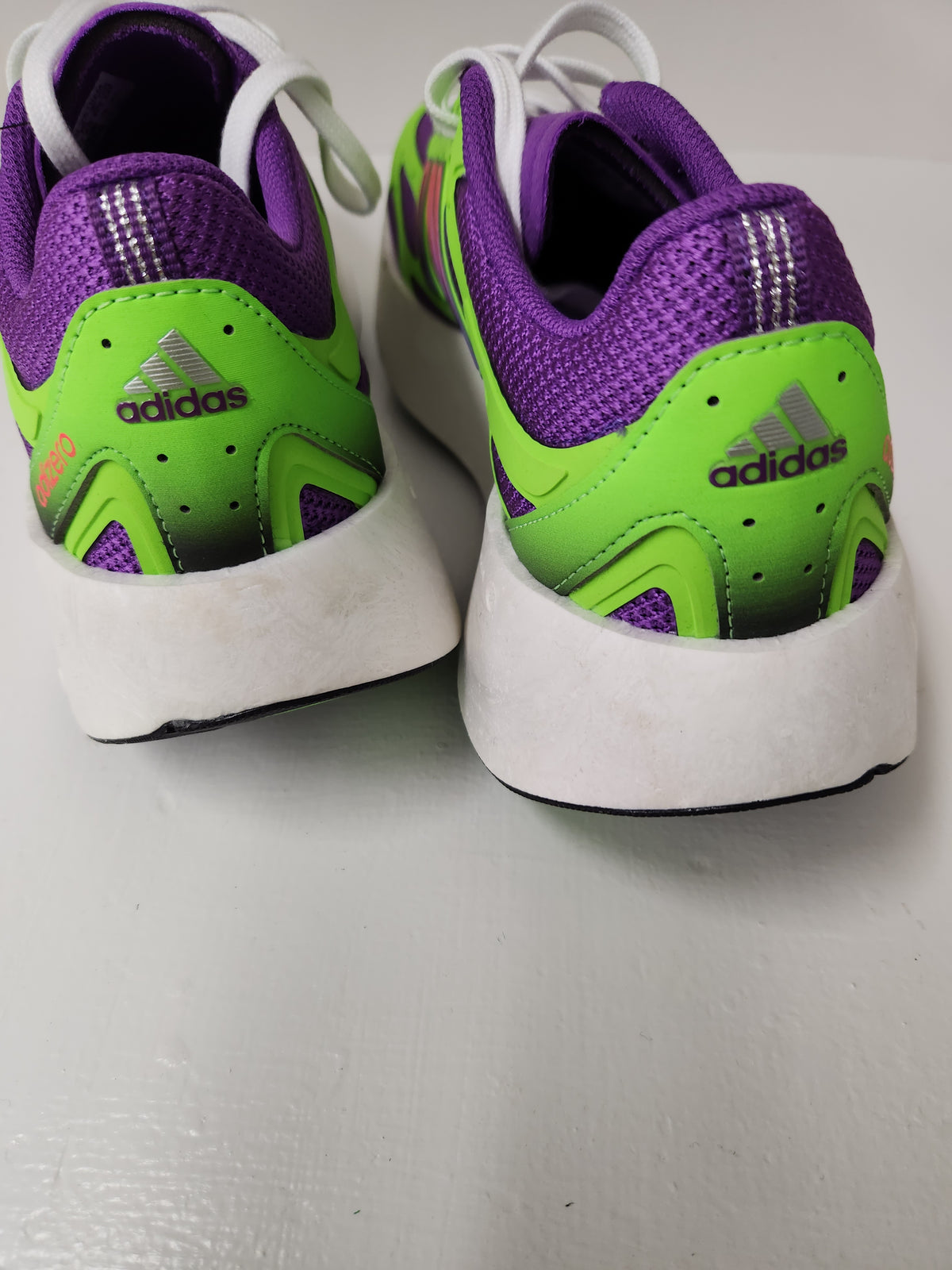 A pair of Adidas running shoes shown from the back, featuring a purple and neon green color scheme with white soles and white laces, resting on a light gray surface.
