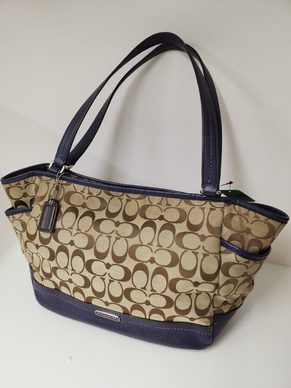 A Coach tote bag in the classic signature monogram canvas, featuring a beige and brown pattern with navy blue leather trim, handles, and base, shown on a plain white background.