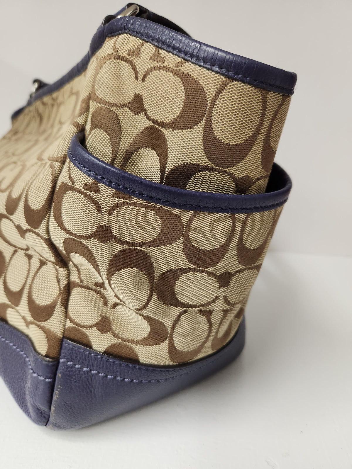 Close-up of a Coach handbag in the signature monogram canvas with navy blue leather trim and base, showing the side pocket detail.
