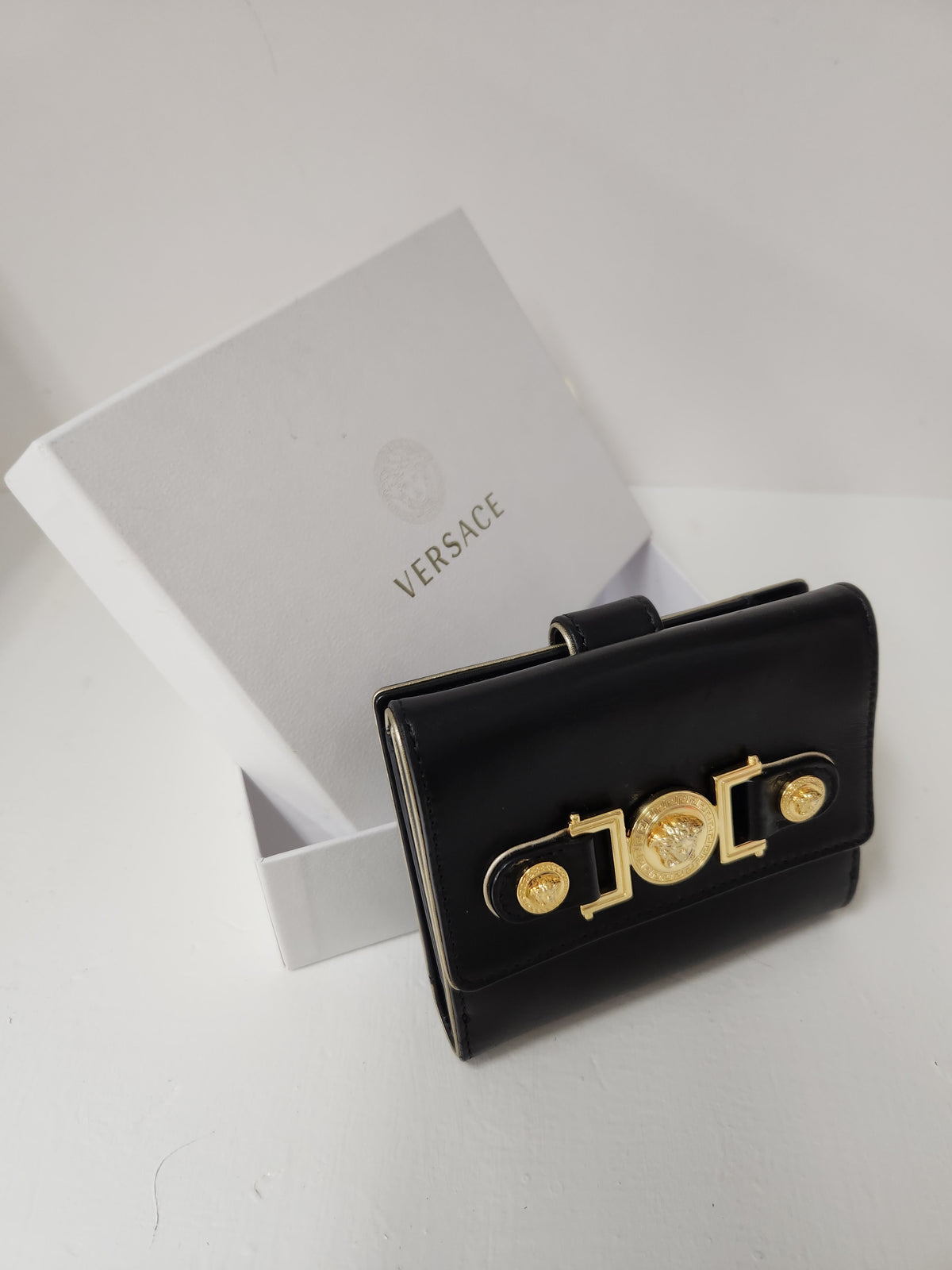 A black leather Versace wallet with a gold-tone Versace logo clasp, shown next to its white branded presentation box on a light gray surface.