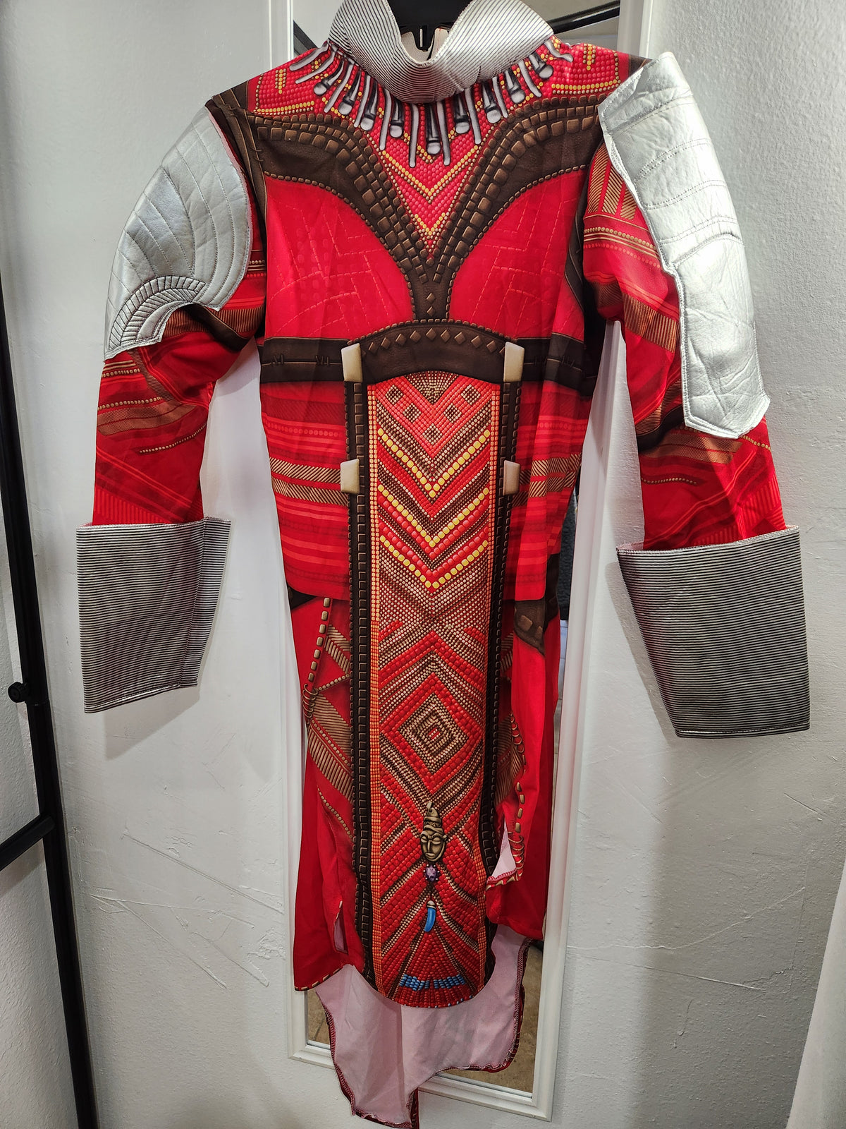 A red and brown patterned costume or tunic with white padded shoulder sections and gray cuffs, displayed against a white wall. Adult Costume