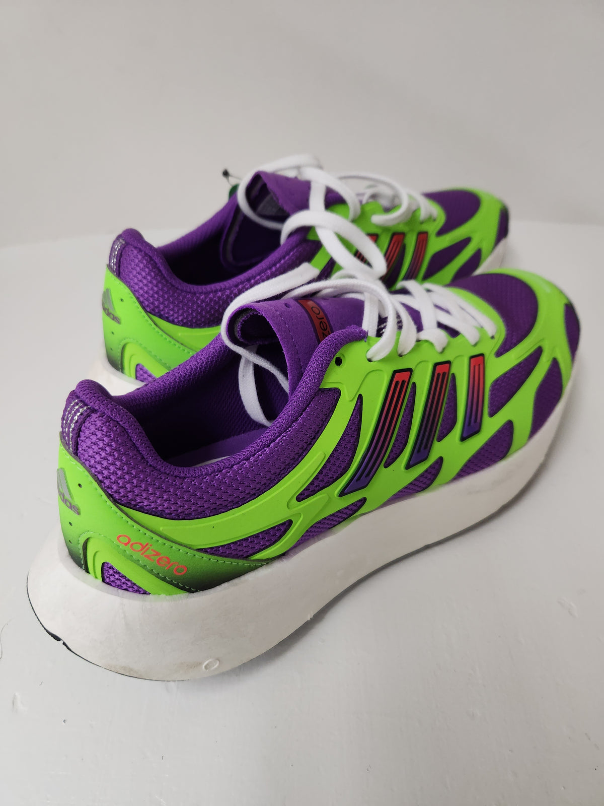 A pair of Adidas running shoes in purple and neon green with white laces and a white sole, photographed on a light gray surface.