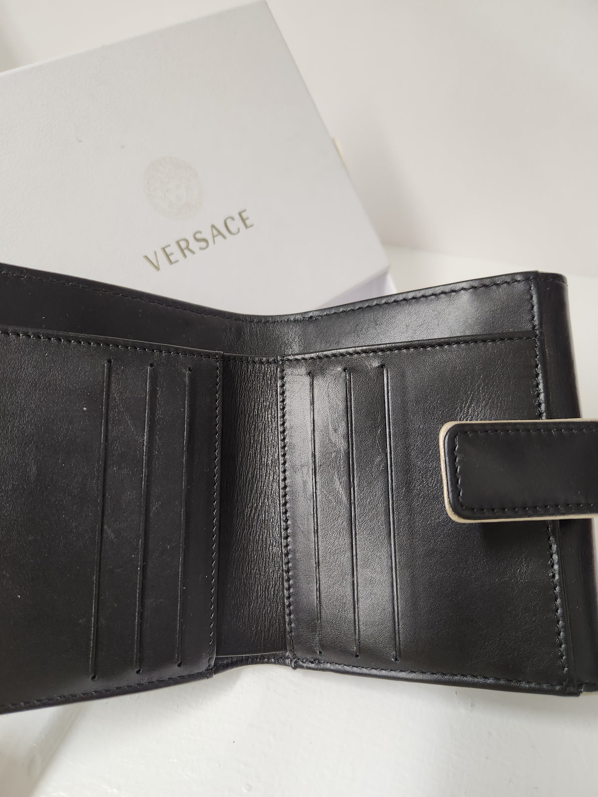 An open black leather bifold wallet with multiple card slots and a small gold-tone metal accent, shown next to a white Versace box on a white surface.