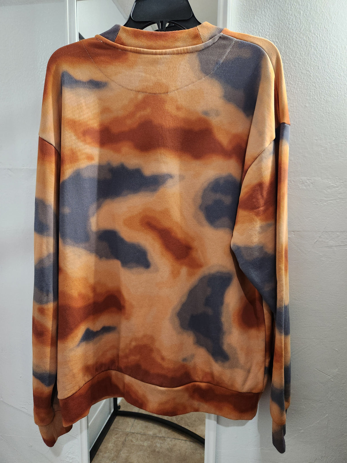 A long-sleeve crewneck sweatshirt with an abstract tie-dye or watercolor pattern in orange, brown, and dark gray, hanging on a black hanger against a white wall.