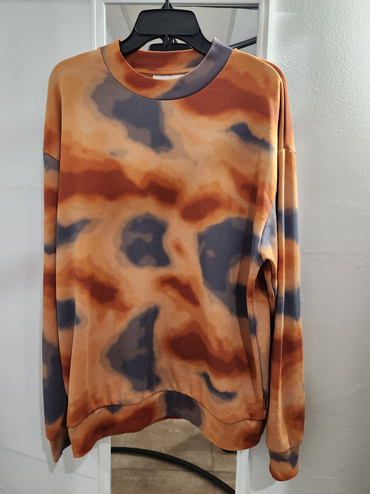 A long-sleeve crewneck sweatshirt with an abstract tie-dye pattern in orange, brown, and dark gray, hanging on a black hanger against a white wall.